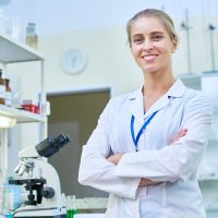 Portrait of young female scientist smiling cheerfully and looking at camera while posing in modern laboratory with arms crossed, copy space