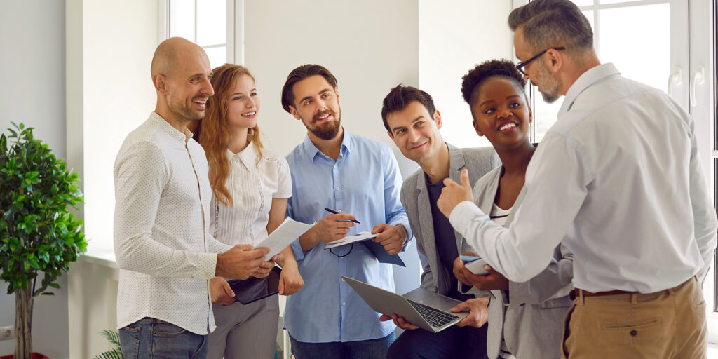 Friendly male business leader or trainer is talking to group of people standing in office near him